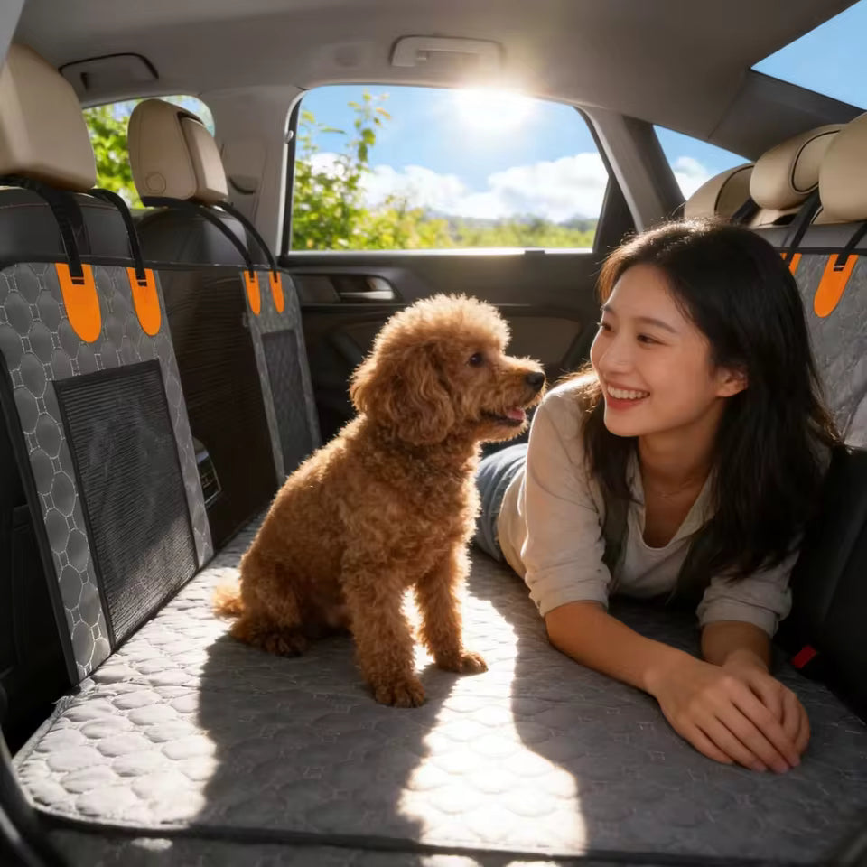 A smiling woman and fluffy brown poodle lounging on a grey quilted car seat cover in the backseat.