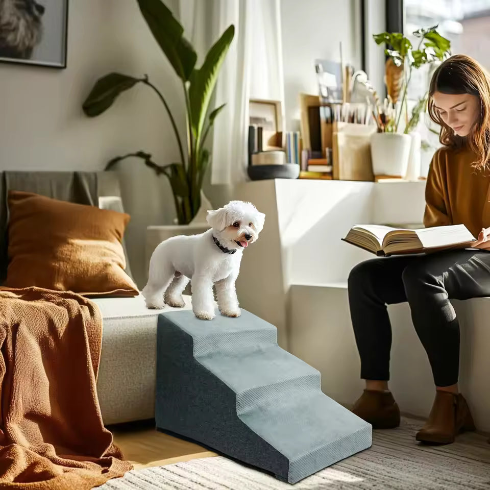 A small white dog stands on a grey 4-step pet stair next to a sofa while a woman reads.