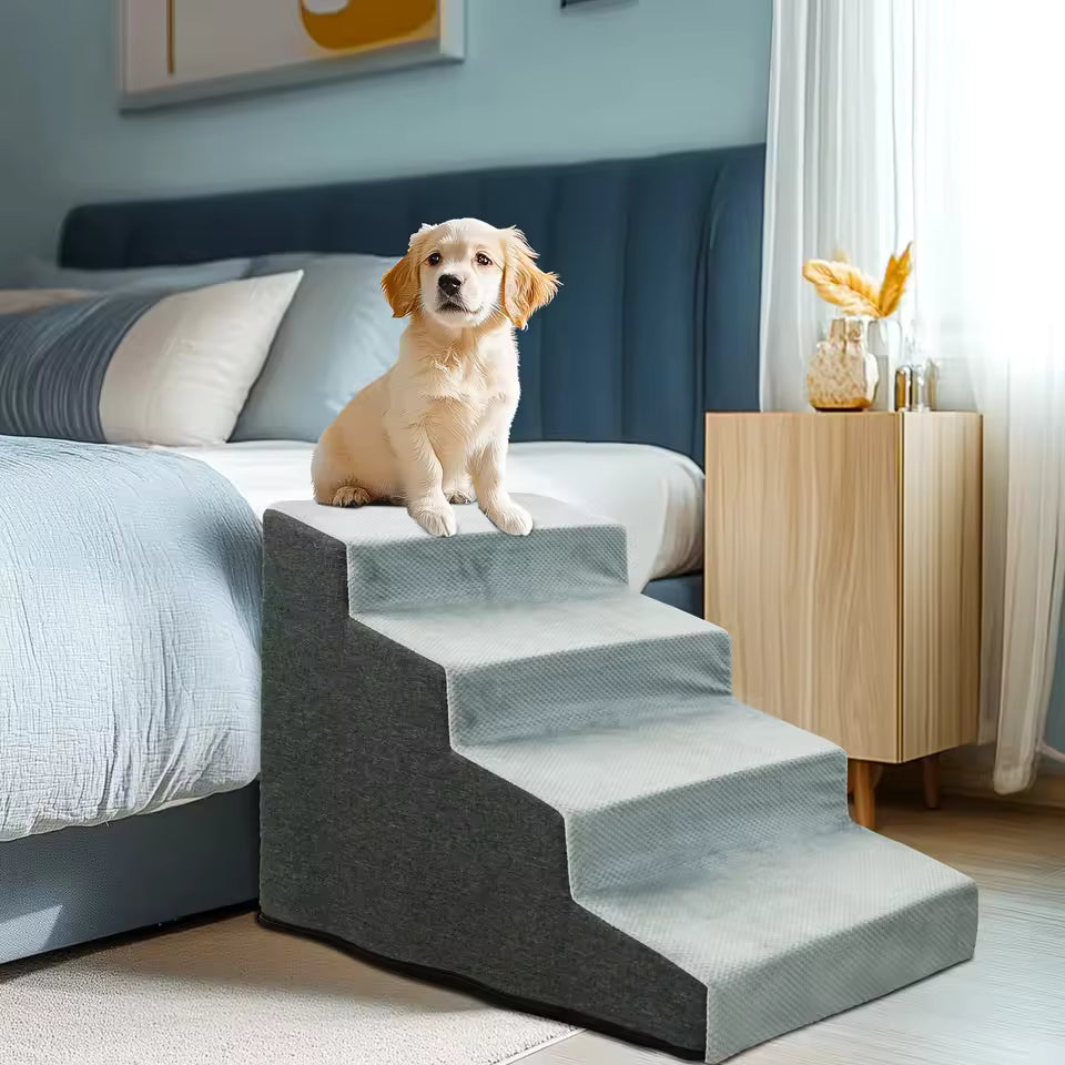 A small puppy sitting atop a four-step grey pet stair next to a bed.