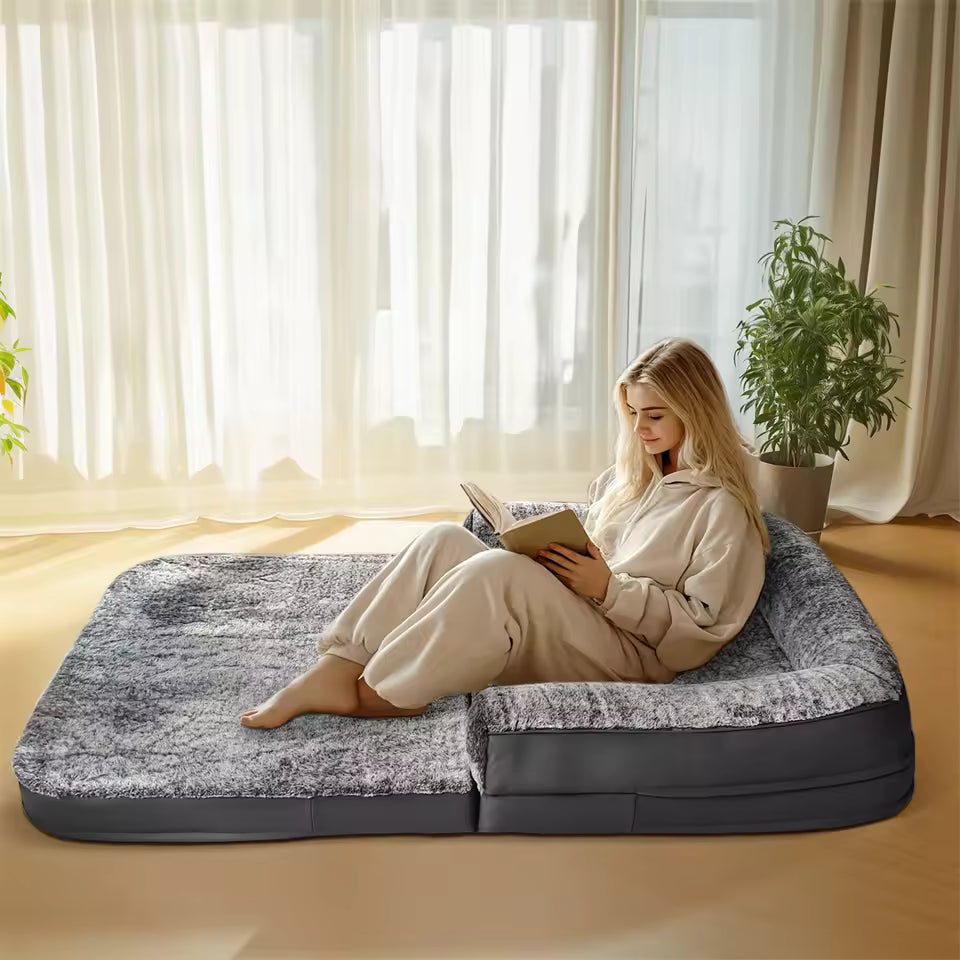 A woman sits and reads a book on a large gray human-sized pet bed in a sunlit room.