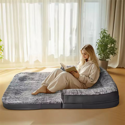 A woman sits and reads a book on a large gray human-sized pet bed in a sunlit room.