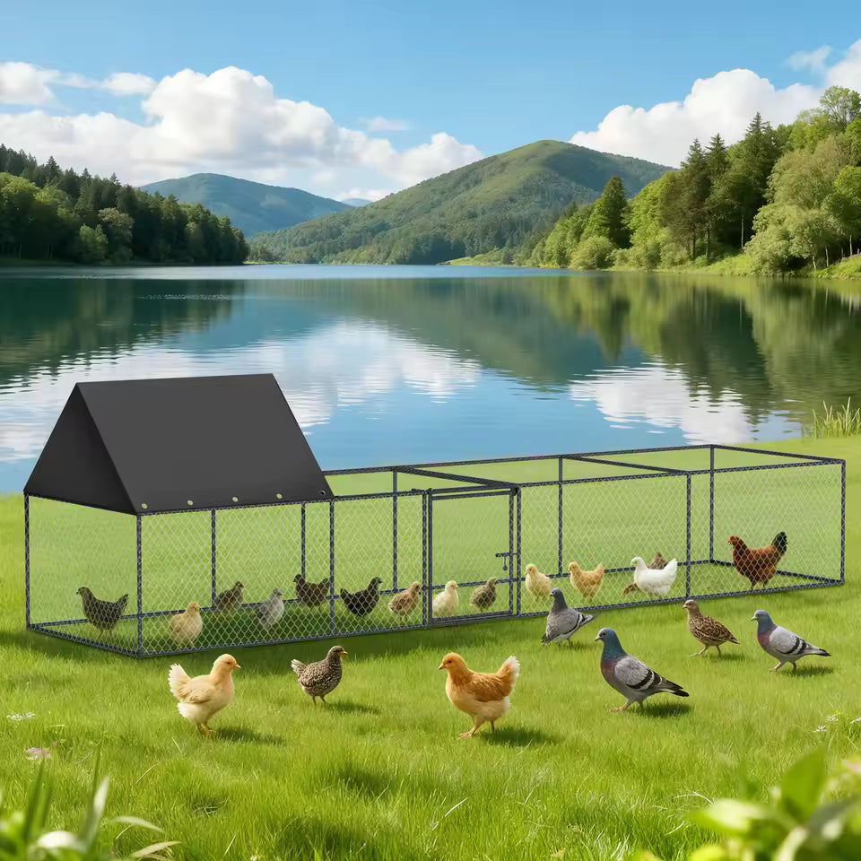 A metal wire chicken coop with a black roof sitting on a grassy shore by a scenic lake with mountains.